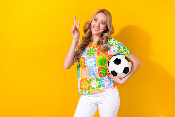 Photo of friendly pretty woman dressed flower print t-shirt showing v-sign playing football empty space isolated yellow color background. - Foto, Imagem