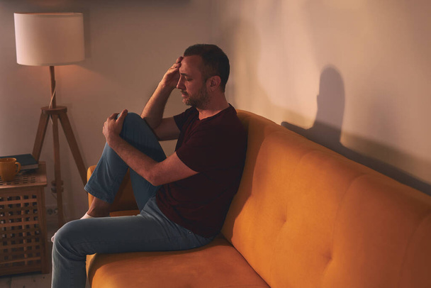 Hombre con dolor de cabeza y dolor de rodilla sentado en un sofá en casa. - Foto, imagen