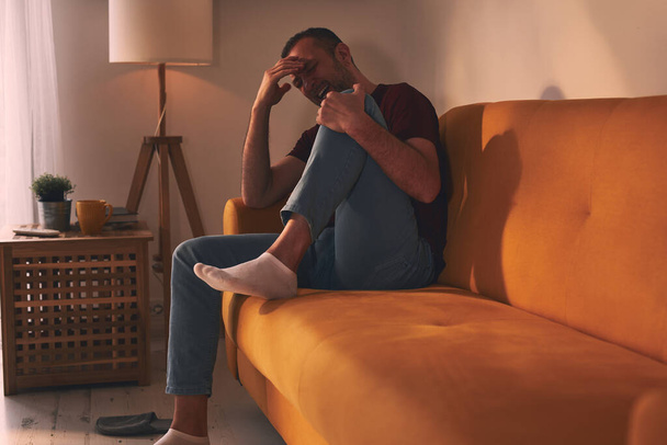 Άνδρας με πόνο στο γόνατο κάθεται σε έναν καναπέ στο σπίτι. - Φωτογραφία, εικόνα