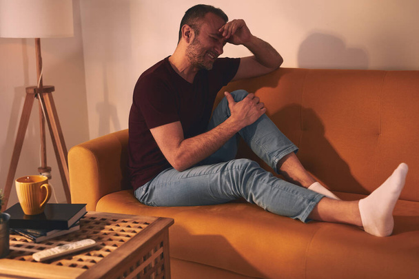 Άνδρας με πόνο στο γόνατο κάθεται σε έναν καναπέ στο σπίτι. - Φωτογραφία, εικόνα