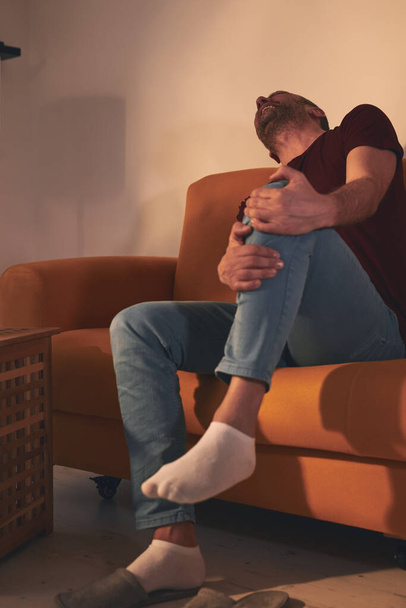 Человек с болью в колене сидит дома на диване. - Фото, изображение