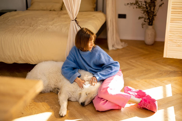 Jeune femme prend soin de son chien blanc mignon tout en étant couché ensemble sur le sol à la maison. Loisirs et amitié avec chien concept - Photo, image