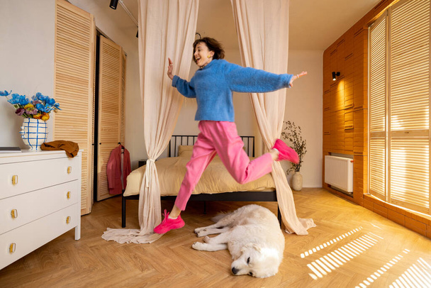 Femme saute par-dessus un chien endormi, s'amusant à passer du temps libre à la maison. Style de vie actif domestique et amitié avec les animaux domestiques - Photo, image