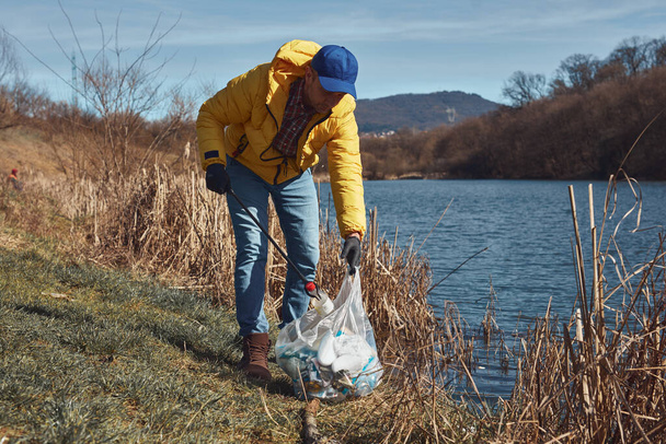 Vrijwilliger en milieuactivist schoonmaken vuile oever meer gevuld met afval. - Foto, afbeelding