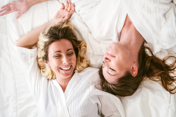 Ευτυχισμένες δύο φίλες Καυκάσιες νεαρές γυναίκες με λευκά μπουρνούζια να απολαμβάνουν και να κοιτάζονται μαζί με χαμόγελα ξαπλωμένες στο κρεβάτι. ΛΟΑΤΚΙ ζευγάρια διασκέδαση δραστηριότητες αναψυχής spa ομορφιά φυσική θεραπεία - Φωτογραφία, εικόνα
