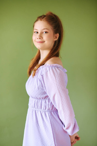 Beauty portrait of pretty young 15 - 16 year old redhaired teeenage girl wearing purple dress posing on green background - Photo, Image