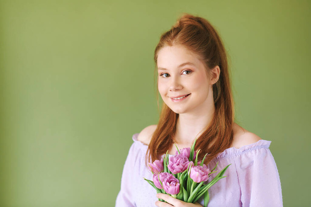 Beauty portrait of pretty young 15 - 16 year old redhaired teeenage girl wearing purple dress posing on green background, holding tulip flowers - Photo, Image