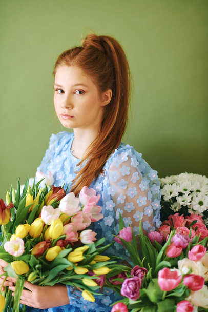 Portrait of pretty young 15 - 16 year old red-haired girl surrounded by many spring flowers posing on green background, beauty and fashion concept - Φωτογραφία, εικόνα