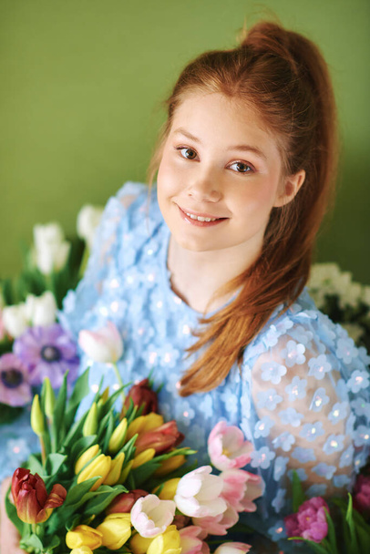 Portrait of pretty young 15 - 16 year old red-haired girl surrounded by many spring flowers posing on green background, beauty and fashion concept - Foto, imagen