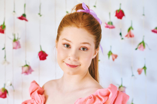 15-16 yaşlarında, pembe mercan elbise giyen, beyaz arka planda asılı çiçekler, güzellik ve moda konseptiyle poz veren güzel bir genç kızın stüdyo portresi. - Fotoğraf, Görsel