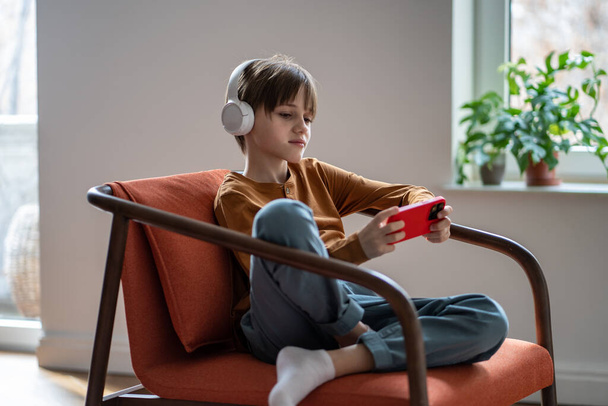 Rustige jongen in hoofdtelefoon ontspannen op de fauteuil gebruik telefoon als video game console. Geïnteresseerde kleine schooljongen rusten Bekijk video podcast op mobiele telefoon scherm online in headset. Verslaving van kinderen - Foto, afbeelding