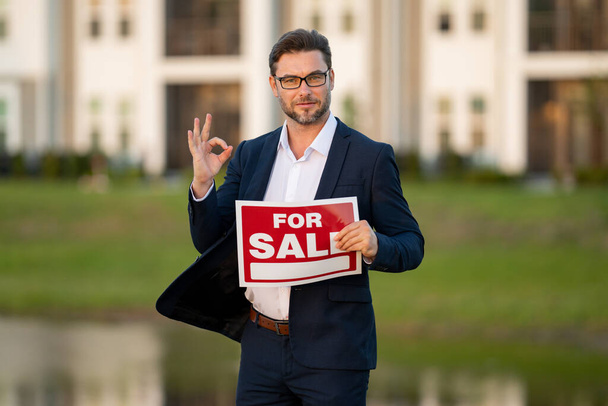 Handsome real estate agent holding banner house for sale. House with land and insurance. Business man real estate agent in business suit presenting the house for sale. Land agent trying to sell house - Photo, image