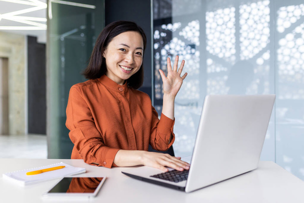 Portrait of young successful business woman in modern office using laptop, Asian woman with smiling and looking at camera, holding hand up in greeting gesture - Foto, immagini