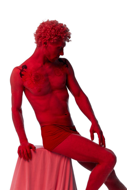 Atractivo modelo masculino sentado en la caja que cubre la tela y posando sin camisa en un filtro rojo aislado sobre un estudio blanco como una estatua. Concepto de estética corporal, belleza masculina, creatividad, inspiración, arte - Foto, Imagen