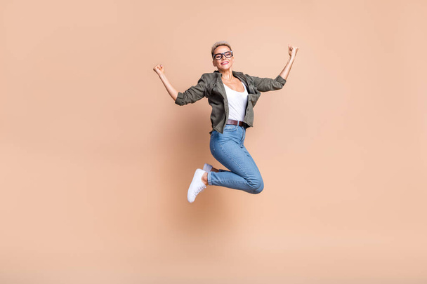 Full length photo of excited lucky lady dressed khaki shirt jumping high rising fists isolated beige color background. - Φωτογραφία, εικόνα