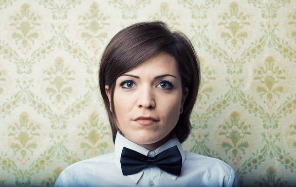 Young woman with boyish hair, sharp frontal portrait. Vintage wallpaper backdrop, white shirt, beautiful, large eyes. Serious, intelligent, and self-assured with an elegant bowtie. Calm and confident  - Photo, Image
