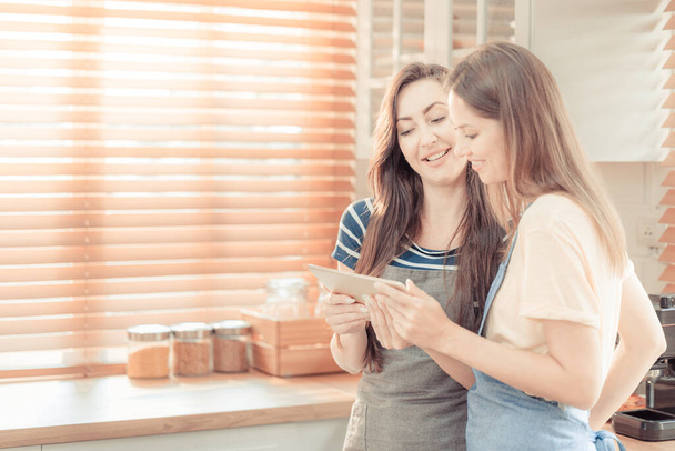 Beautiful lesbian couple cooking healthy food, looking on digital tablet at modern kitchen interior. Homosexual relations and healthy eating concept.  LGBTQ+,LGBT,LGBTQ home online kitchen. warm tone. - Foto, afbeelding