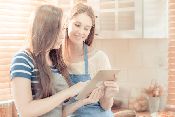 Beautiful lesbian couple cooking healthy food, looking on digital tablet at modern kitchen interior. Homosexual relations and healthy eating concept.  LGBTQ+,LGBT,LGBTQ home online kitchen. warm tone. - Fotó, kép