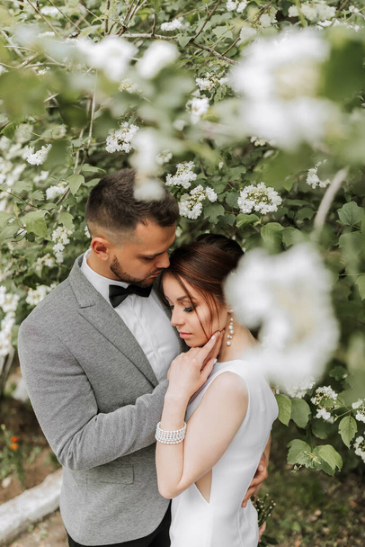 the bride in a white evening dress poses with the groom with a beautiful hairstyle, the groom gently wraps the bride and leans against her against the background of spring flowers. - Φωτογραφία, εικόνα