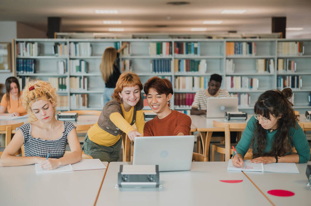 Vrolijke jonge multiraciale vrienden glimlachen en wijzen naar laptop scherm tijdens de voorbereiding van examens in de buurt van geconcentreerde mannelijke en vrouwelijke klasgenoten in de moderne bibliotheek - Foto, afbeelding