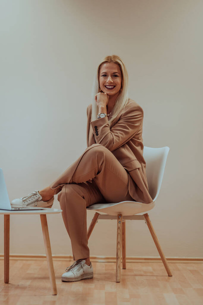 A professional businesswoman sits on a chair, surrounded by a serene beige background, diligently working on her laptop, showcasing dedication and focus in her pursuit of success. - Valokuva, kuva