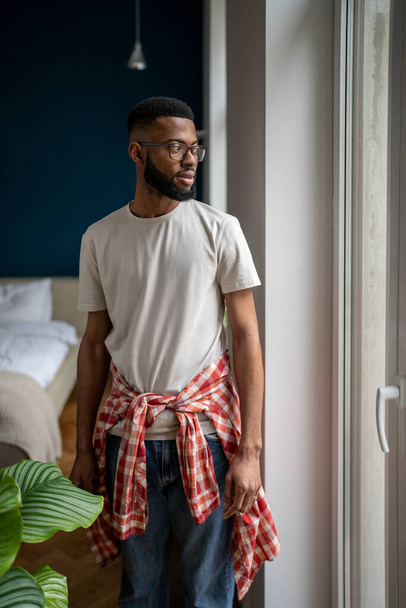 Domowy portret beztroskiego, brodatego Afroamerykanina w okularach, ubranego w stylowe ubranie patrzącego przez okno, właśnie przeniósł się do nowego mieszkania. Ludzie w domu - Zdjęcie, obraz