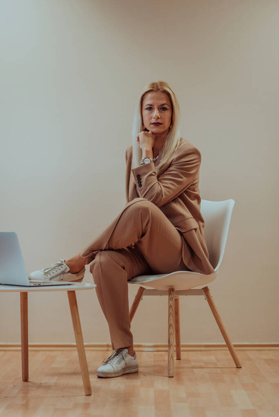 A professional businesswoman sits on a chair, surrounded by a serene beige background, diligently working on her laptop, showcasing dedication and focus in her pursuit of success. - Photo, image