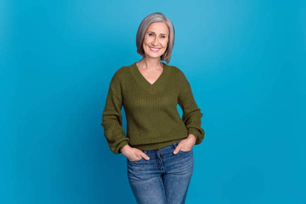 Photo portrait of attractive cheerful senior woman hold hands jeans pants pockets wear khaki sweater isolated blue color background. - 写真・画像