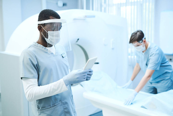 Giovane radiografo afro-americano in maschera e guanti utilizzando tablet in camera MRI mentre il suo collega mettere lenzuolo sul tavolo - Foto, immagini
