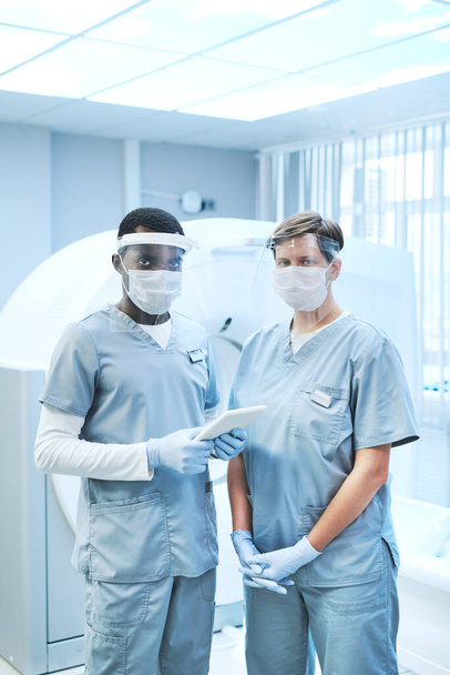 Portrait of interracial nurses in scrubs and protection shields standing against MRI scanner in clinic - Fotografie, Obrázek