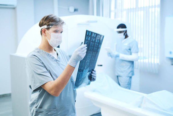 Radiologista focado em escudo de segurança trabalhando com imagem de raio-X do cérebro contra tomógrafo no hospital - Foto, Imagem