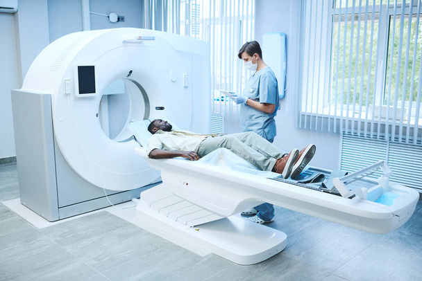 MRI specialist in facial mask and gloves standing at patient lying on table and filling his card online - Foto, afbeelding