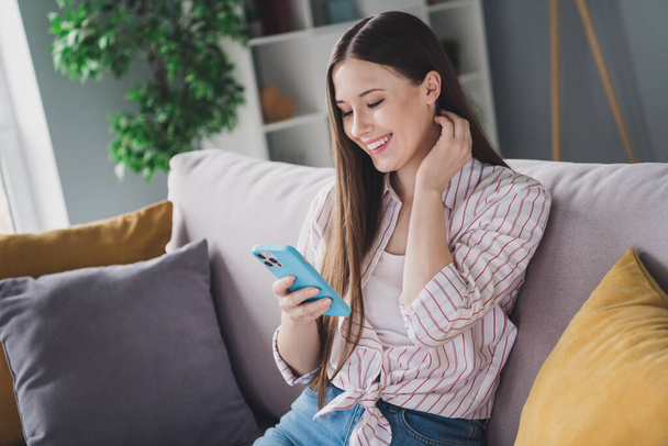 Çizgili tişörtlü, sevimli genç bir bayanın koltukta oturup modern cihazları kapalı ev odasında mesajlaşırken fotoğrafı.. - Fotoğraf, Görsel