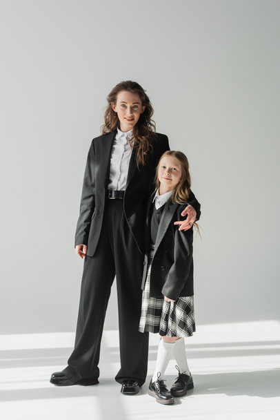 modern mother and daughter, businesswoman in suit hugging schoolgirl in uniform with plaid skirt, on grey background, blazers, getting ready for new school year, looking at camera, formal attire  - Fotografie, Obrázek