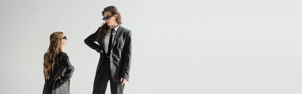 modern mother and daughter in sunglasses, businesswoman in suit and schoolgirl in uniform looking at each other while standing on grey background in studio, fashionable family, banner  - Photo, Image