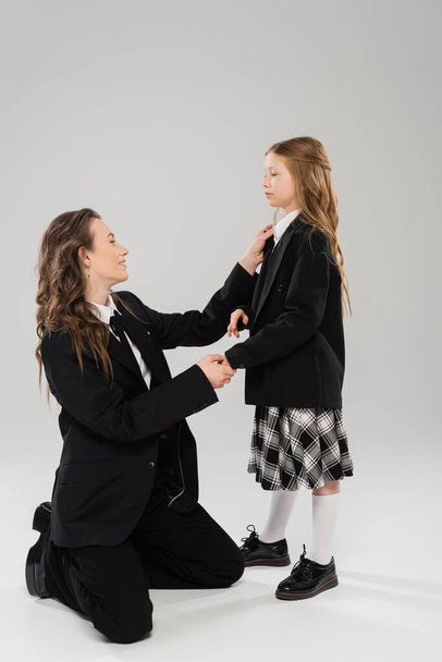 mother adjusting tie of schoolgirl, happy woman in business attire and daughter on grey background in studio, back to school, getting ready, school preparation, modern parenting, parent-child  - Photo, Image