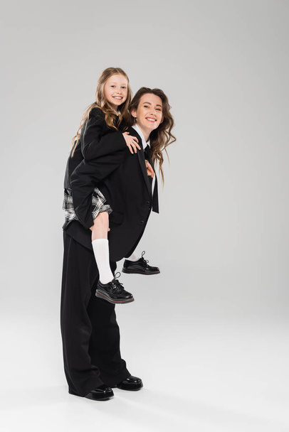 cheerful mother piggybacking her daughter, working mom in business attire and schoolgirl in uniform on grey background in studio, modern parenting, fashionable family, having fun  - Photo, Image