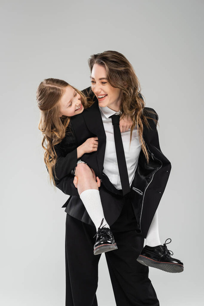 mother and daughter having fun, cheerful woman piggybacking schoolgirl, working mom in formal attire and kid in uniform on grey background in studio, modern parenting  - Foto, afbeelding