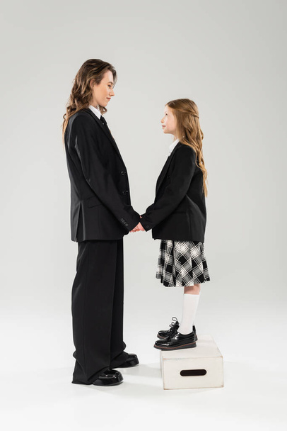 mother and child holding hands, happy woman in business attire and child in school uniform standing on step stool on grey background, modern parenting, face to face - Photo, Image