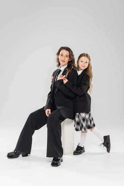 happy girl hugging working mother, businesswoman sitting on concrete stool near kid in uniform on grey background in studio, formal attire, parent-child, modern parenting, fashion shoot  - Fotografie, Obrázek