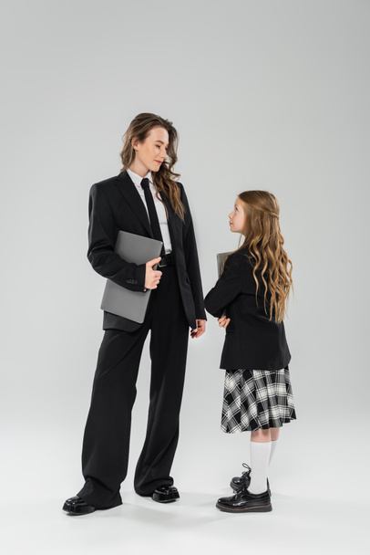 working mother and daughter, digital nomadism, remote work, e learning, businesswoman in suit and schoolgirl standing together with laptops on grey background in studio, modern parenting  - Photo, Image