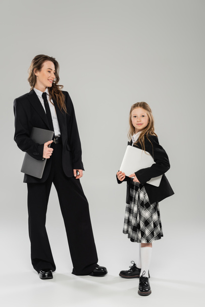 mother and daughter, digital nomadism, remote work, e learning, happy businesswoman in suit and schoolgirl standing together with laptops on grey background in studio, modern parenting  - Photo, Image