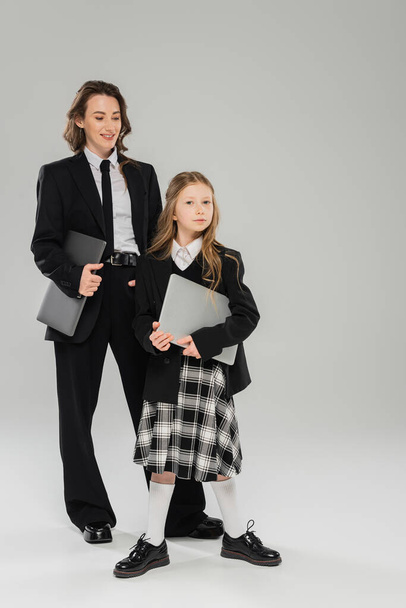happy mother and daughter, digital nomadism, remote work, e learning, businesswoman in suit and schoolgirl standing together with laptops on grey background in studio, modern parenting  - Photo, Image