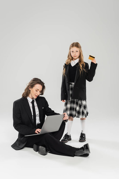 working mother and daughter, digital nomadism, schoolgirl holding credit card near mom using laptop on grey background, modern parenting, business attire, financial education, online purchase - Photo, Image