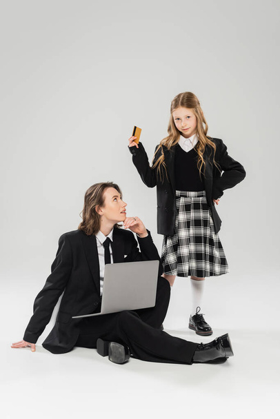 mother and schoolgirl, digital nomadism, girl holding credit card near working mom using laptop on grey background in studio, modern parenting, business attire, online purchase, e-commerce  - Photo, Image