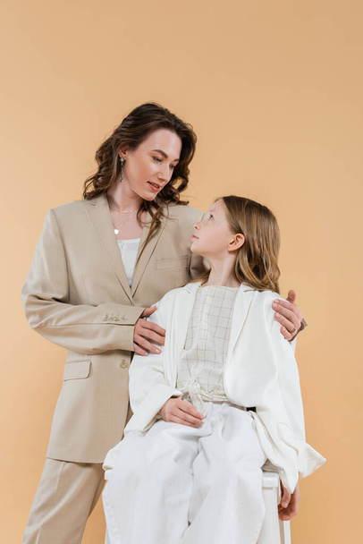 üzleti anya és lánya öltönyben, nő ölelés vállát lány ül széken bézs háttér, divatos ruhák, hivatalos öltözék, vállalati anya, modern család  - Fotó, kép