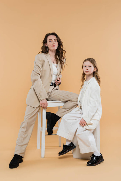 corporate mom and daughter in suits, woman sitting on high chair and looking at camera near girl on beige background, fashionable outfits, formal attire, corporate mom, motherhood and career  - Photo, Image
