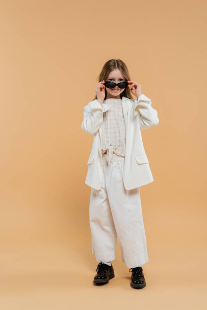 trendy preteen girl in white suit and black shoes looking at camera while wearing sunglasses and standing on beige background, fashionable outfit, formal attire, child model, trendsetter, style  - Foto, Bild