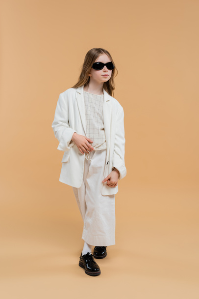 trendy preteen girl in white suit, sunglasses and black shoes posing and standing on beige background, fashionable outfit, formal attire, child model, trendsetter, style, fashionista  - Foto, Bild