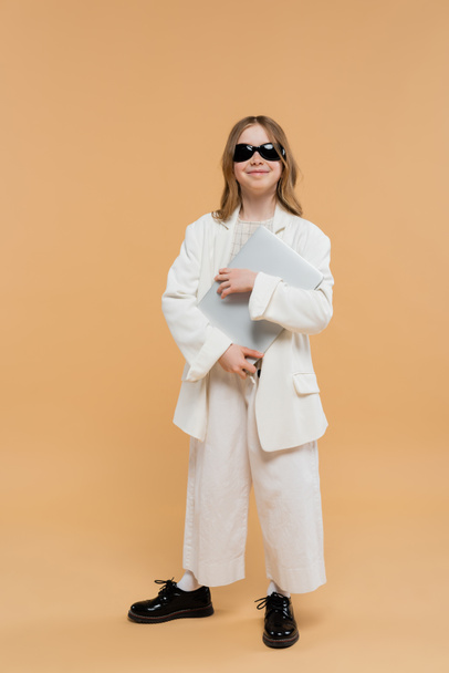 online education, trendy preteen girl in white suit, happy kid in sunglasses holding laptop and standing on beige background, fashionable outfit, formal attire, child model, trendsetter, e-learning  - Foto, Imagem